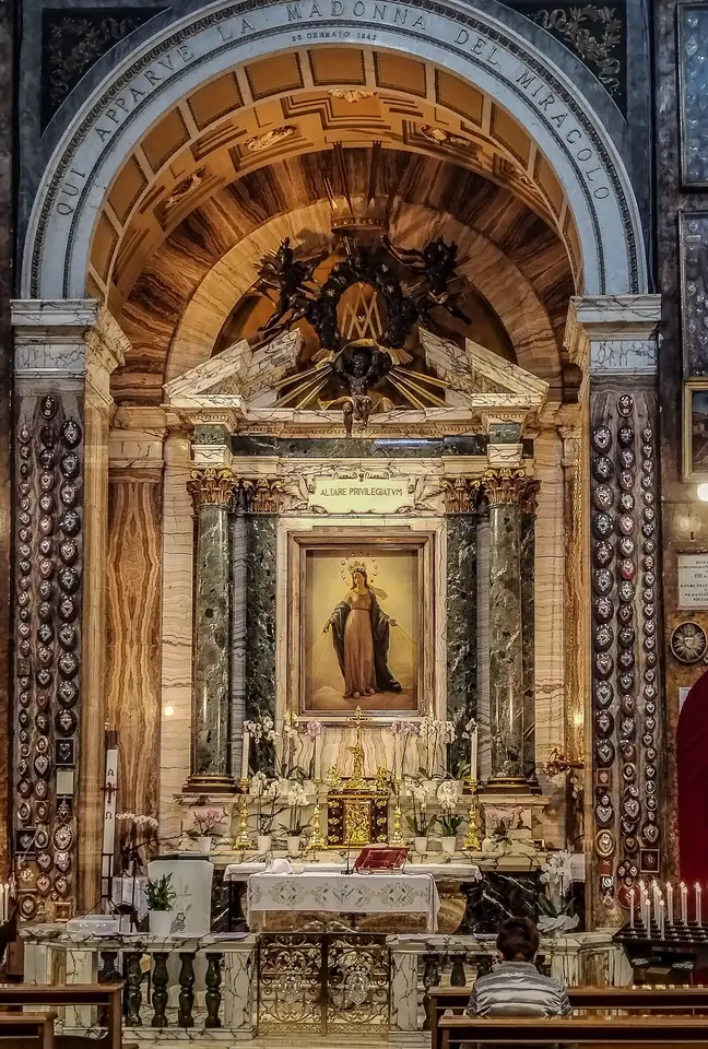 Marble side chapel with a gilded altar, its walls covered with silver votive hearts, around a painting of the Virgin with her arms lowered and rays from her hands