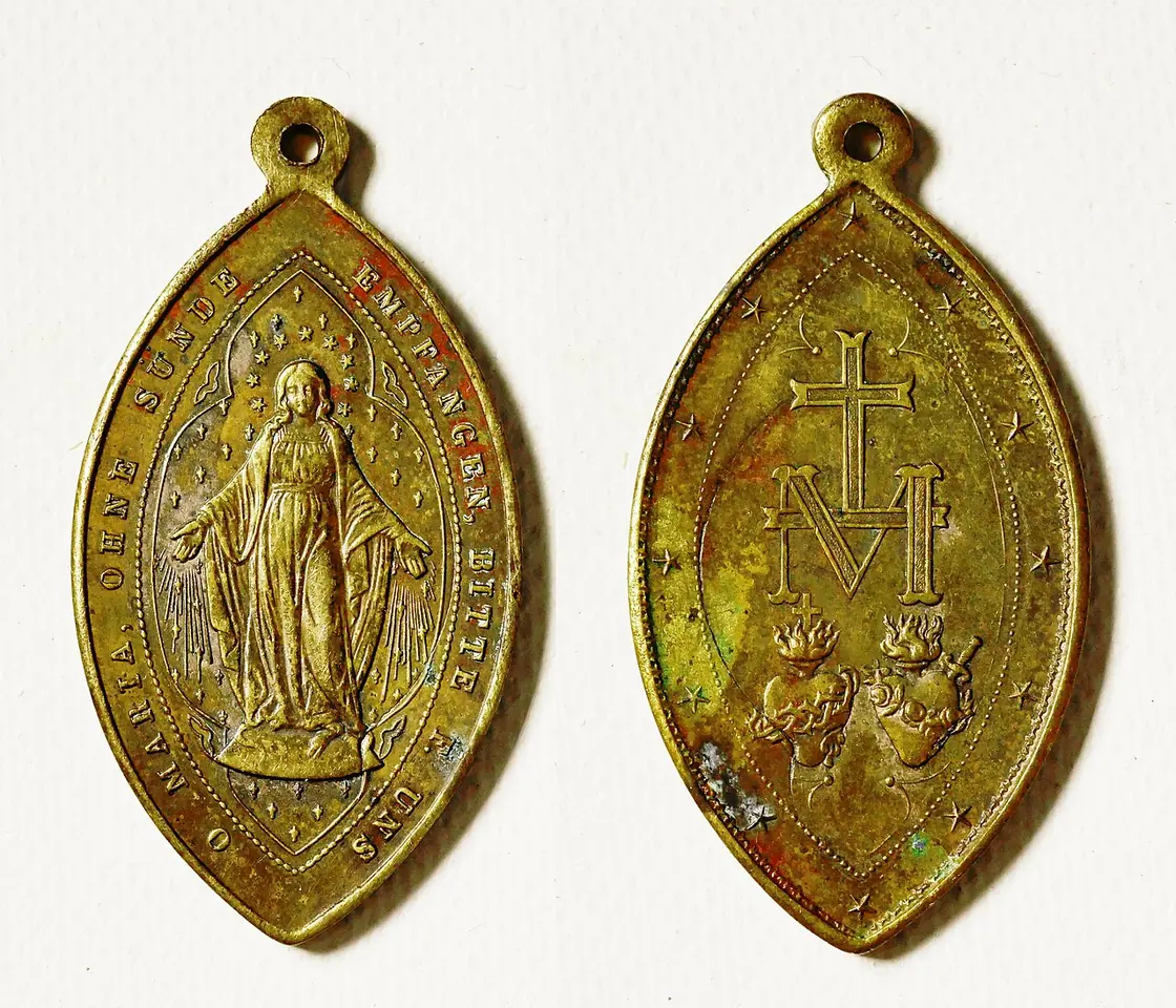 Front and back of a worn brass medal in a pointed oval shape, the Virgin with rays on the front and the M with a cross and two hearts on the back
