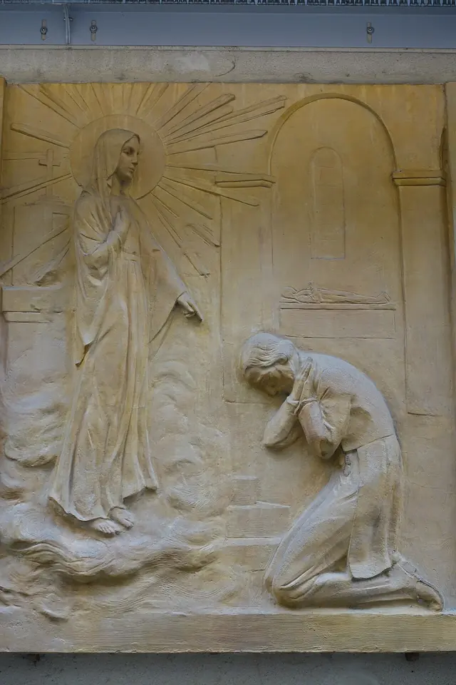 Stone relief of the Virgin Mary standing in a burst of rays as a young nun kneels before her with her face in her hands
