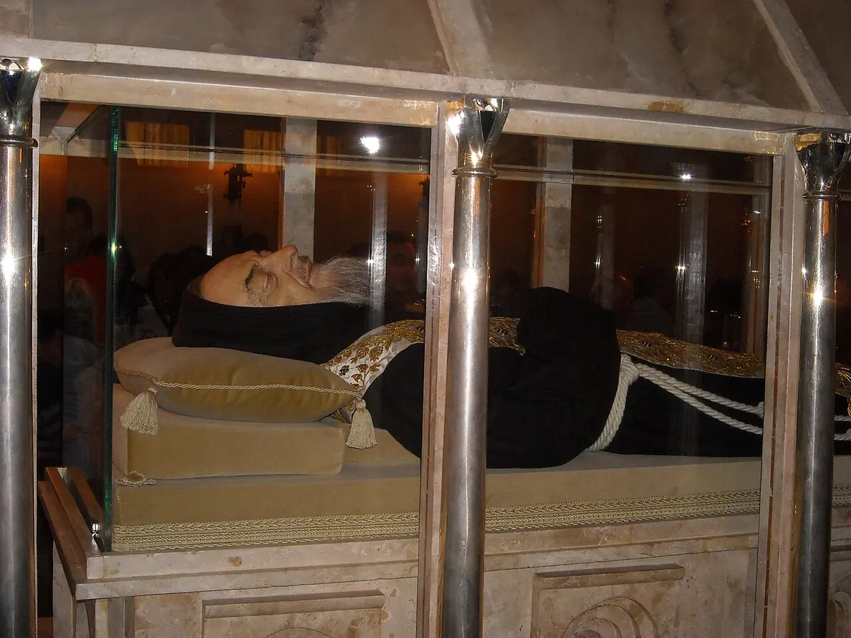 The body of Padre Pio in a brown habit lying in a glass and marble coffin, his face covered by a lifelike mask