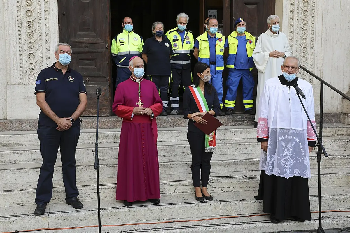 Civil protection volunteers in uniform stand with a bishop, a priest and a mayor on the steps of a church in Rome