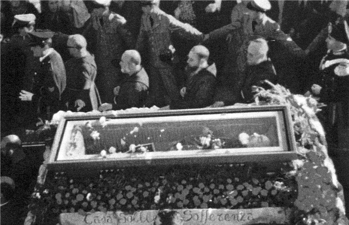 Black-and-white photograph of Padre Pio's coffin with a glass lid, covered in flowers, surrounded by friars and police officers