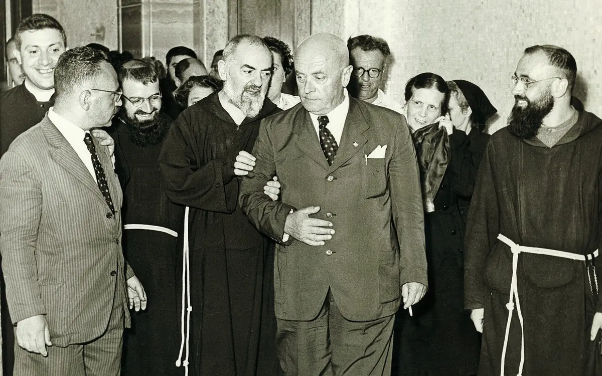 Black-and-white photograph of Padre Pio walking arm in arm with a man in a suit, surrounded by friars and visitors in a corridor