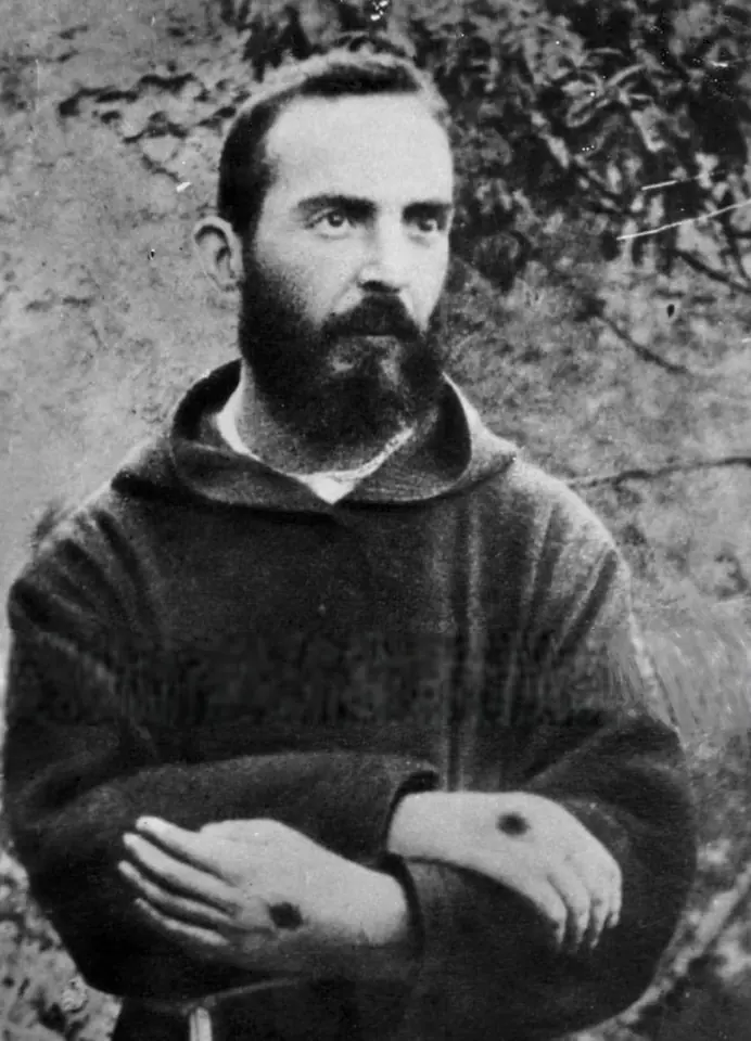 Black-and-white photograph of a young bearded Capuchin friar with dark hair, arms folded, a dark mark visible on the back of one hand