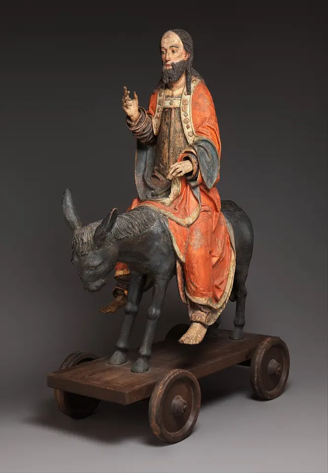 Figura de madera tallada y pintada de Cristo con túnica roja sentado en un asno gris, con la mano derecha alzada para bendecir, sobre una plataforma de madera con ruedas