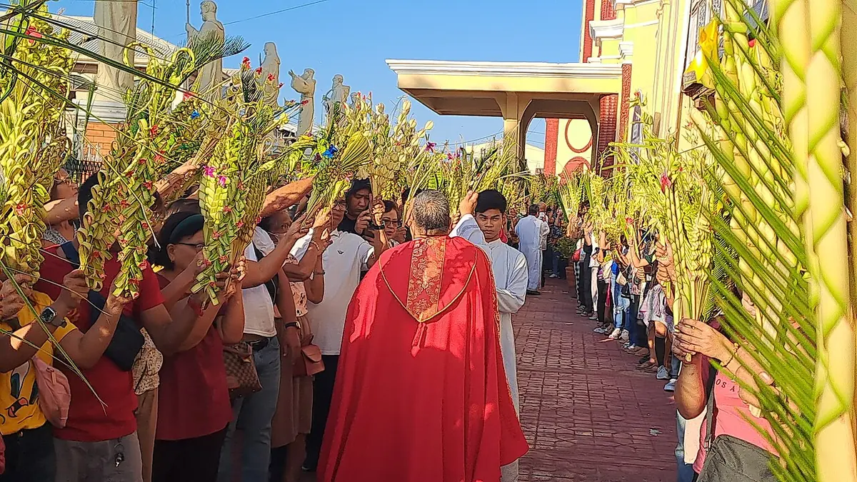 Un sacerdote con capa pluvial roja camina entre dos largas filas de fieles que levantan palmas tejidas mientras las rocía con agua bendita