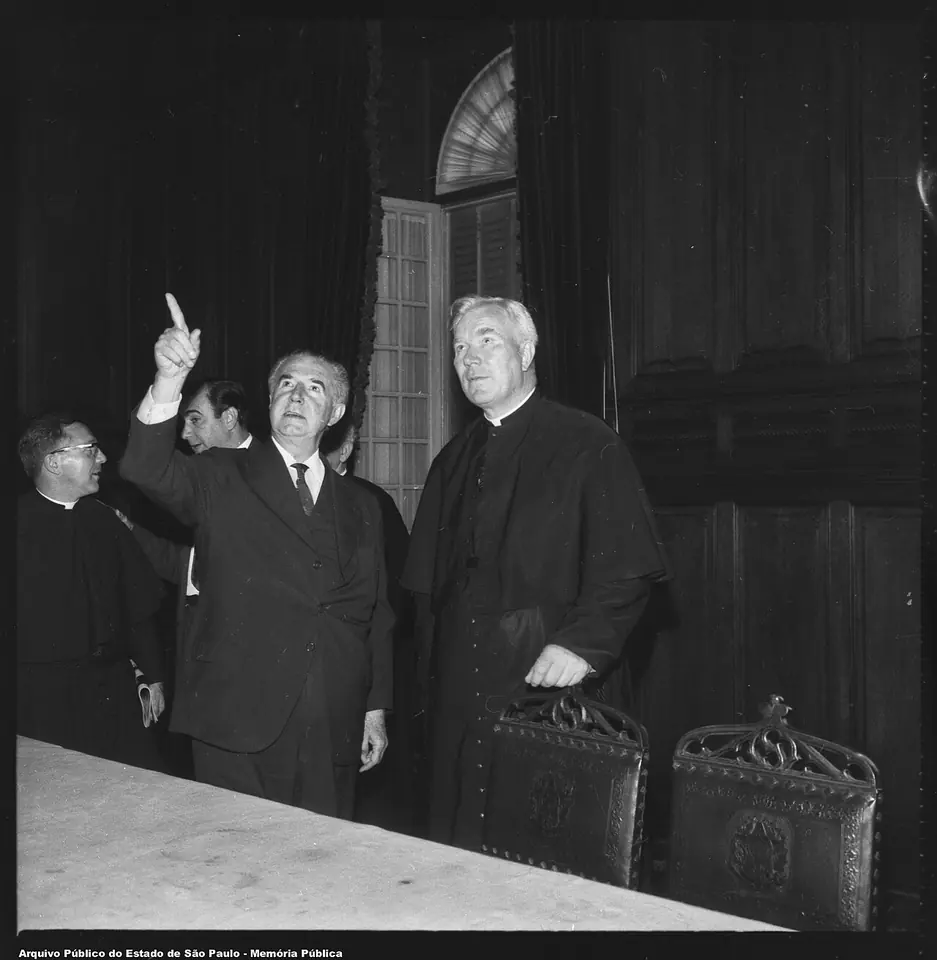 Black-and-white photograph of a tall white-haired priest in a cassock standing beside a man in a dark suit who points upward, with another priest behind them, in an ornate room