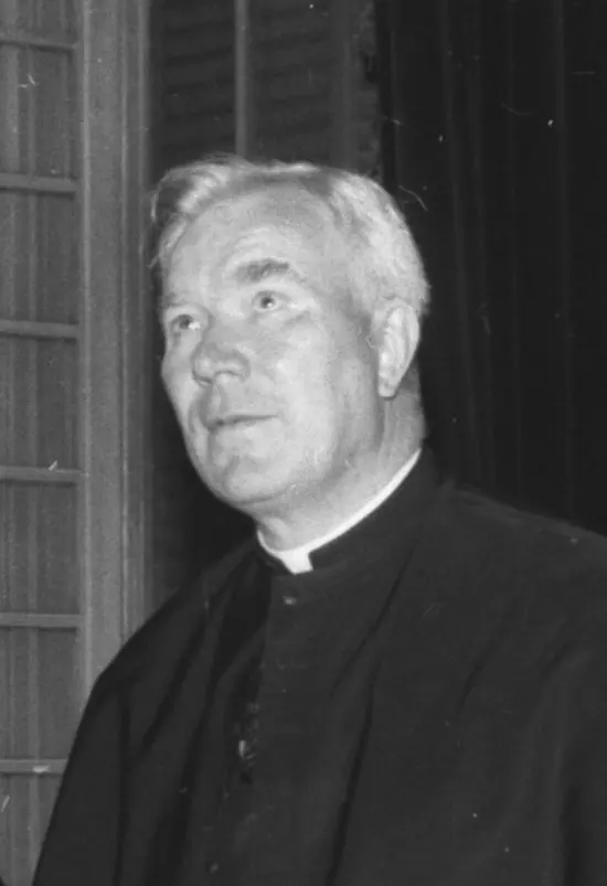 Black-and-white photograph of a white-haired priest in a black cassock and Roman collar, looking up and to the side with a calm, intent expression