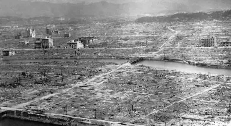 Black-and-white photograph of a flattened city stretching to the hills, with a few gutted concrete buildings standing among the ruins beside a river