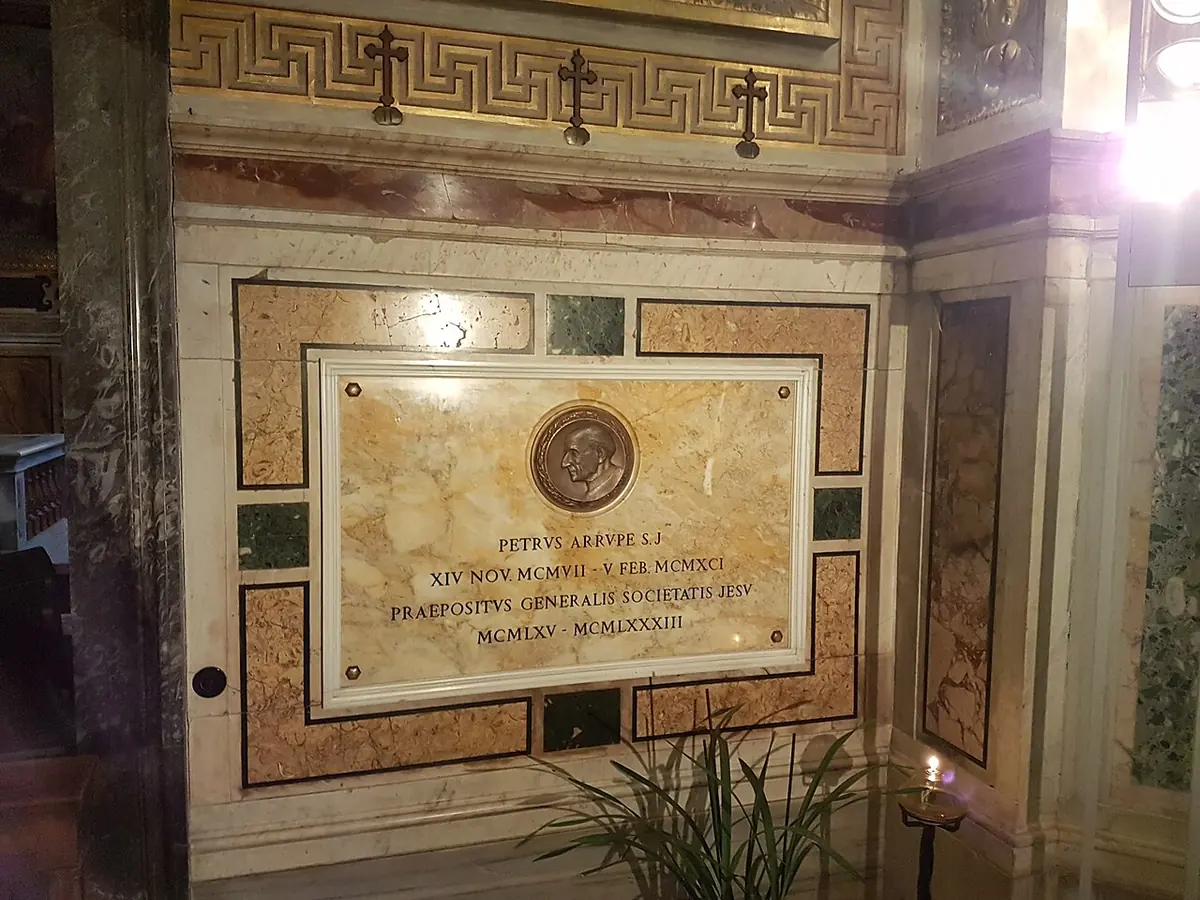 A marble wall slab inside a church with a small bronze medallion and a Latin inscription for Petrus Arrupe, Superior General of the Society of Jesus, with a lit candle beside it