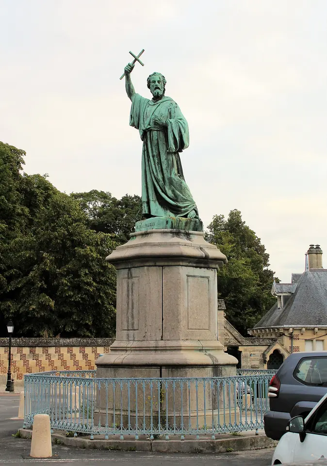 A green bronze statue of a bearded hermit holding a cross high in his right hand, on a tall stone pedestal behind a low iron railing, in a town square