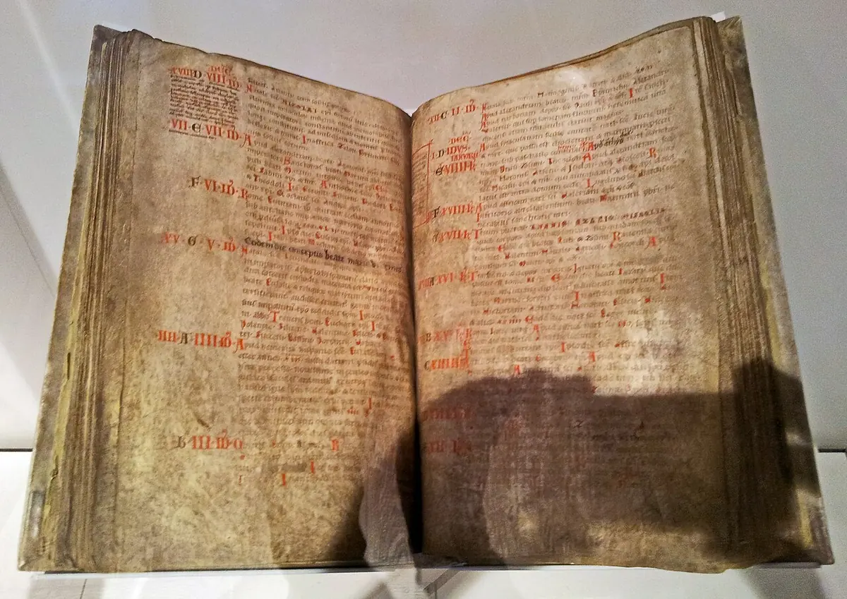 A large open medieval book on display, its worn parchment pages filled with columns of brown writing and red headings