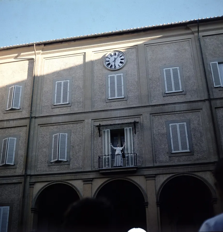 Color photograph of a tall ochre palace with green shutters and a clock, and a small figure in white on the central balcony raising his arms