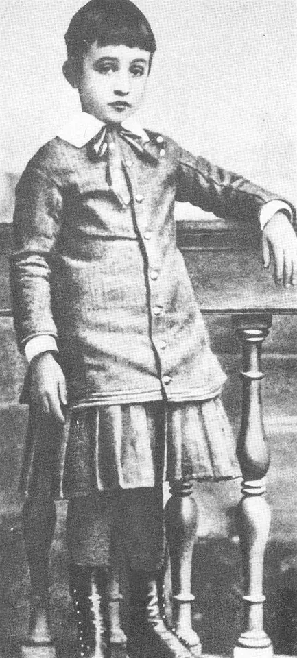 Old full-length photograph of a small boy with short dark hair in a buttoned jacket and knee breeches, leaning on a chair