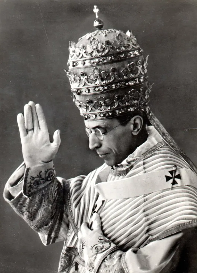 Black-and-white photograph of a pope wearing the triple-crowned papal tiara and embroidered vestments, raising his right hand in blessing