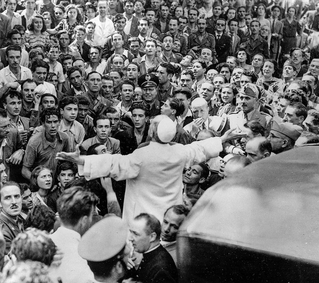 Black-and-white photograph of a pope in white seen from behind with his arms spread wide, surrounded by a dense crowd of men and women reaching toward him