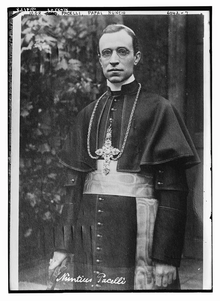 Black-and-white photograph of a tall, thin archbishop in a cassock and short cape with a pectoral cross, standing with his hands folded