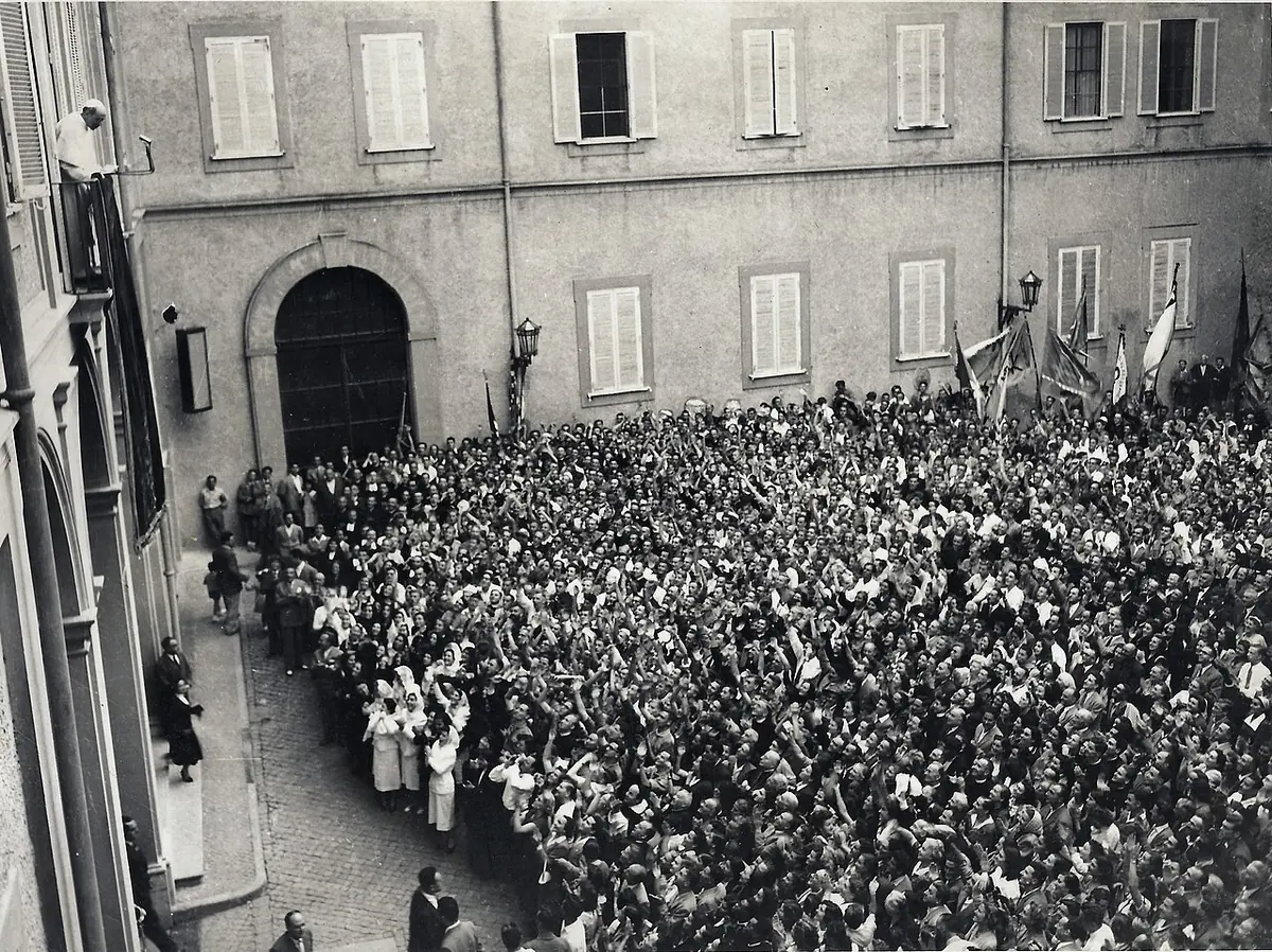 Black-and-white photograph of a large crowd of young pilgrims with banners filling the courtyard of a palace, looking up at a balcony