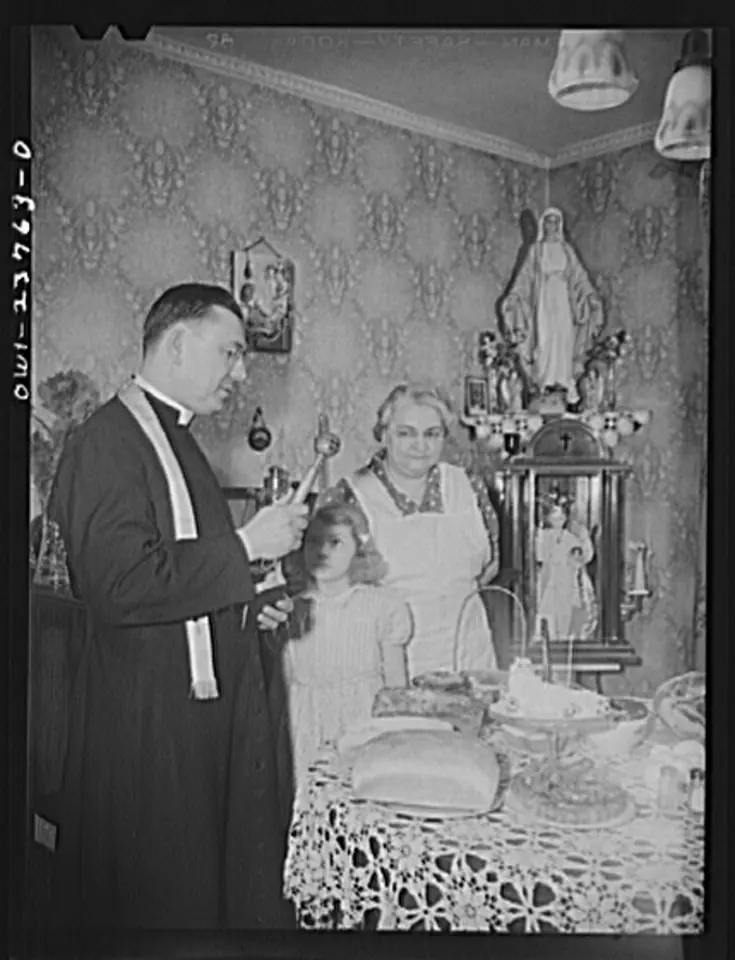 Black-and-white photograph of a priest in a cassock and stole sprinkling holy water over a family's Easter table while a grandmother and a little girl watch, a statue of Our Lady on the shelf behind them