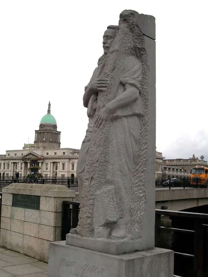 A dark stone statue of Matt Talbot as a thin working man in a long coat, his hands clasped, standing on a plinth beside the River Liffey