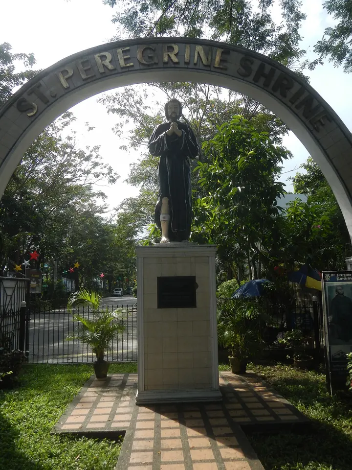 A dark statue of St. Peregrine with folded hands stands on a white pedestal under an arched gateway lettered St. Peregrine Shrine, with trees and a garden path behind