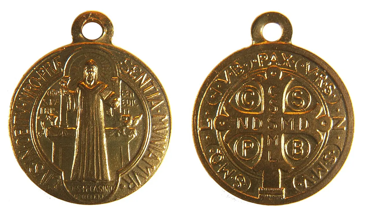The two sides of a small gold St. Benedict medal, one with the standing figure of St. Benedict, the other with a cross covered in Latin initials