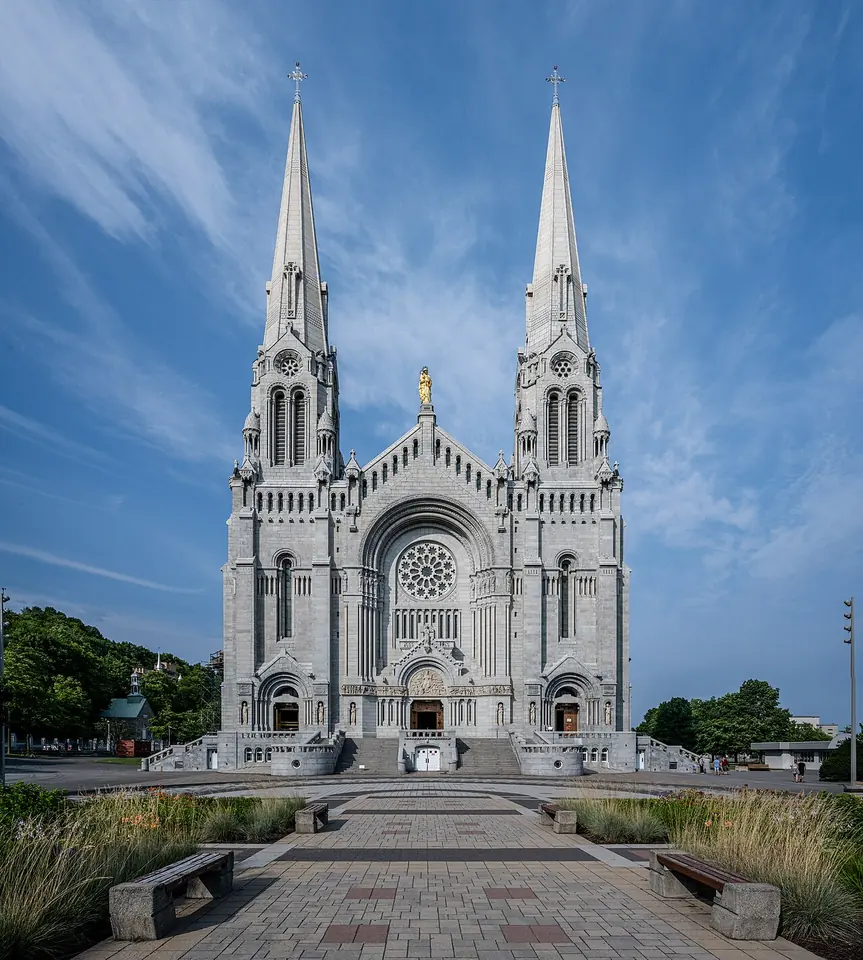 The white stone basilica of Sainte-Anne-de-Beaupré with two tall spires and a gold statue between them, seen across a paved plaza under a blue sky