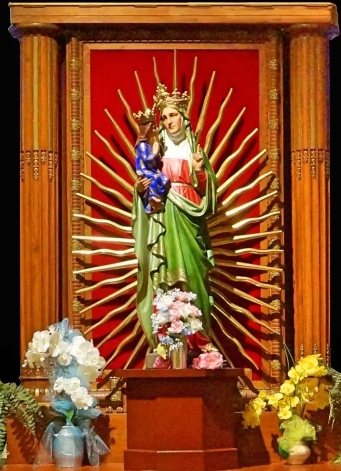 Crowned statue of St. Anne in a green mantle holding the child Mary, set against gilded rays in a red niche with flowers below