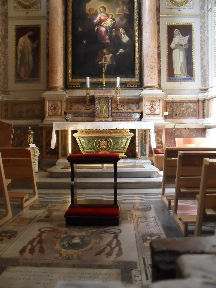 Side chapel of a Roman church with an altar and painting framed by marble columns, a green ancient sarcophagus beneath the altar and a kneeler in front