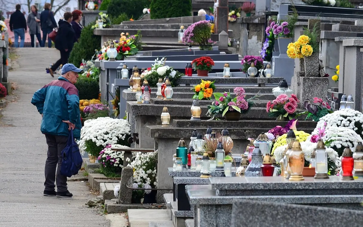 Rows of stone graves in a Polish cemetery covered with glass candle lanterns and pots of white and yellow chrysanthemums, with visitors walking past