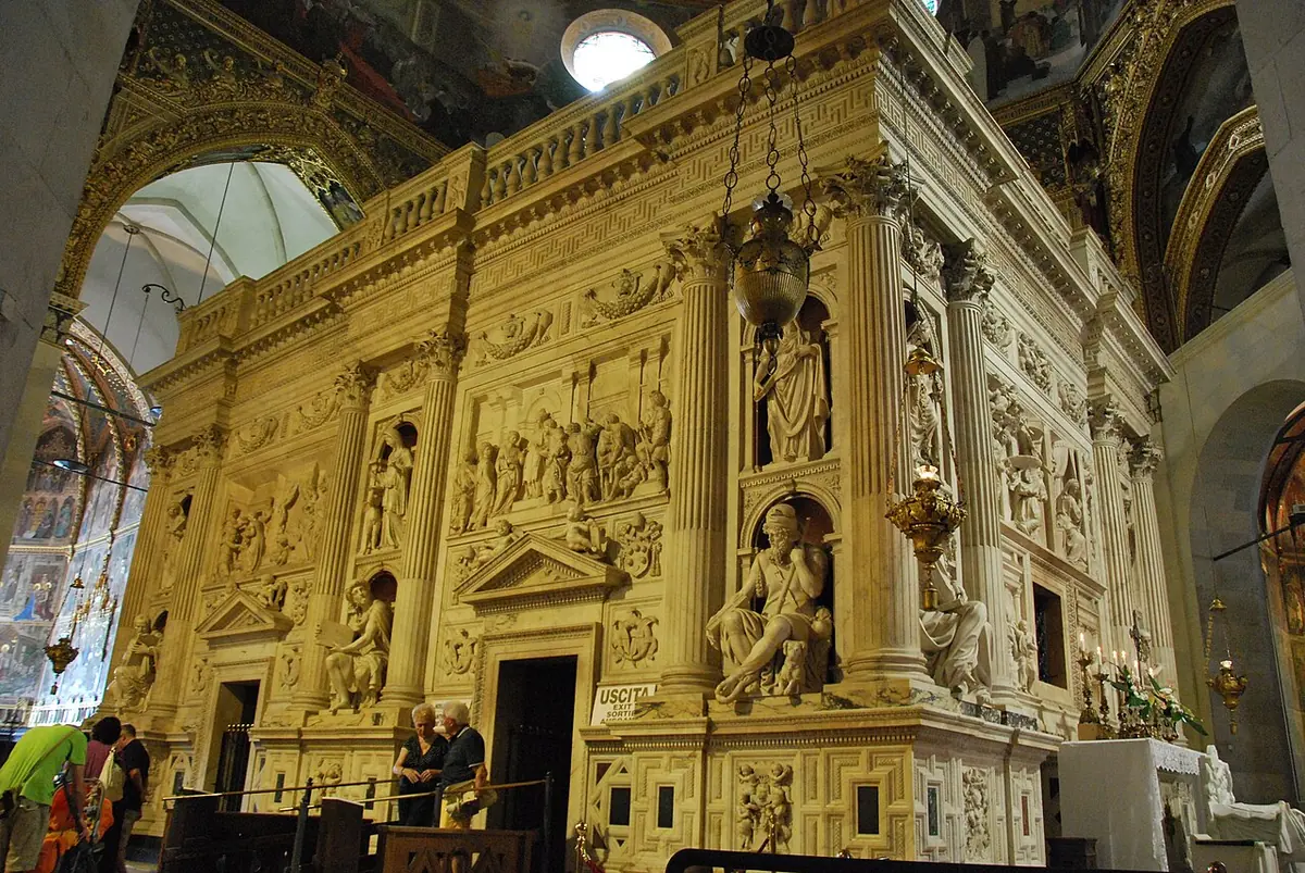 White marble enclosure carved with reliefs and statues in niches, standing inside a large church, with visitors at its doorway