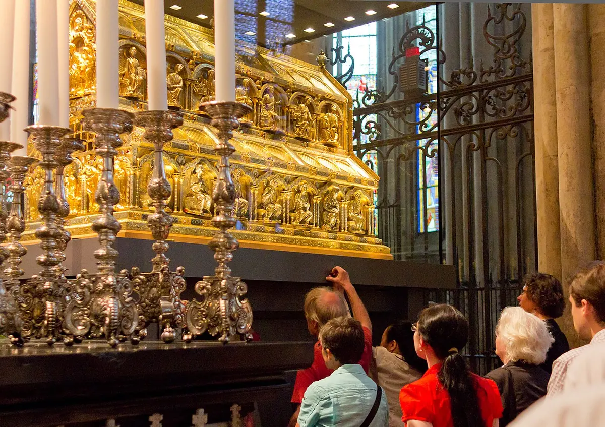 Gilded house-shaped shrine raised on a high stand behind candles, with pilgrims lining up to pass underneath it
