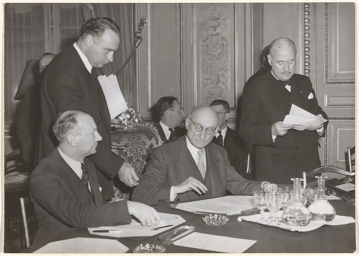 Black-and-white press photograph of Robert Schuman seated at a conference table with papers and water glasses while other diplomats stand and sit around him in an ornate room