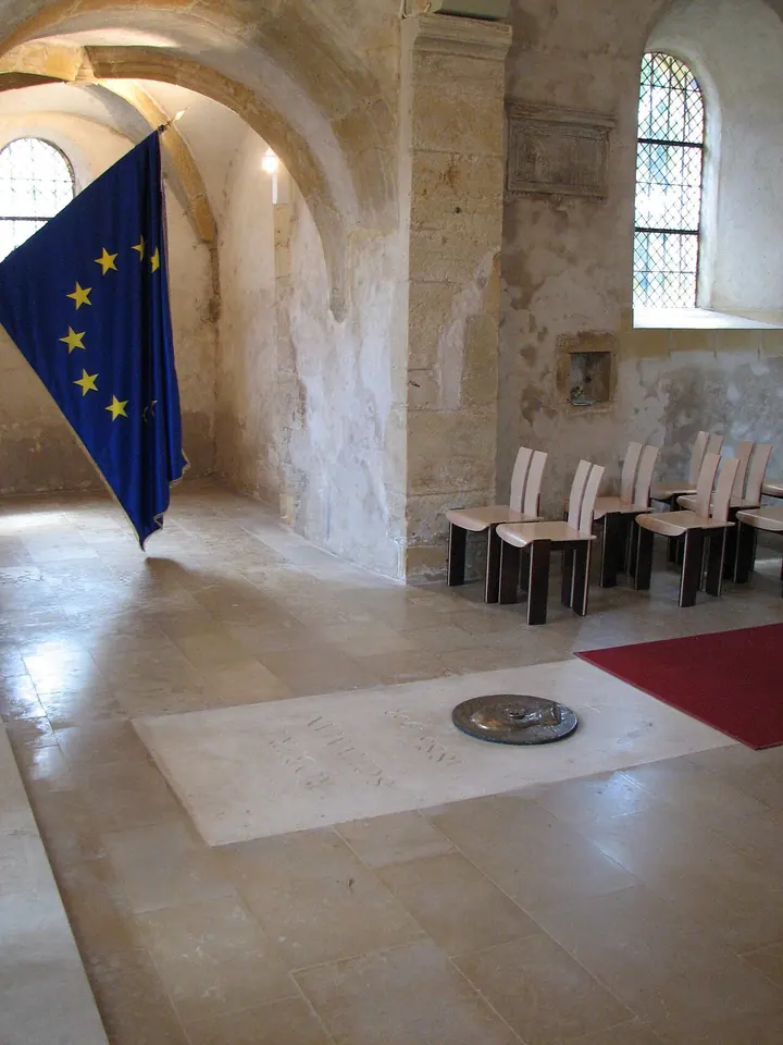 A plain stone slab in the floor of a vaulted church, with a European flag hanging beside it and a row of chairs along the wall