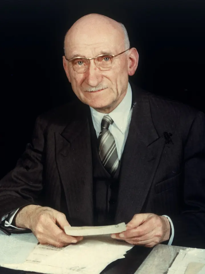 Color photograph of an elderly bald man with round glasses and a gentle smile, in a dark suit and striped tie, seated at a desk holding papers