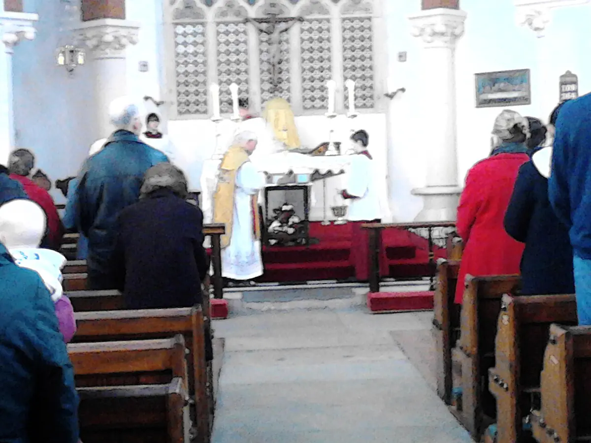View down the aisle of a parish church as a priest in white vestments at the altar blesses candles before the congregation standing in the pews