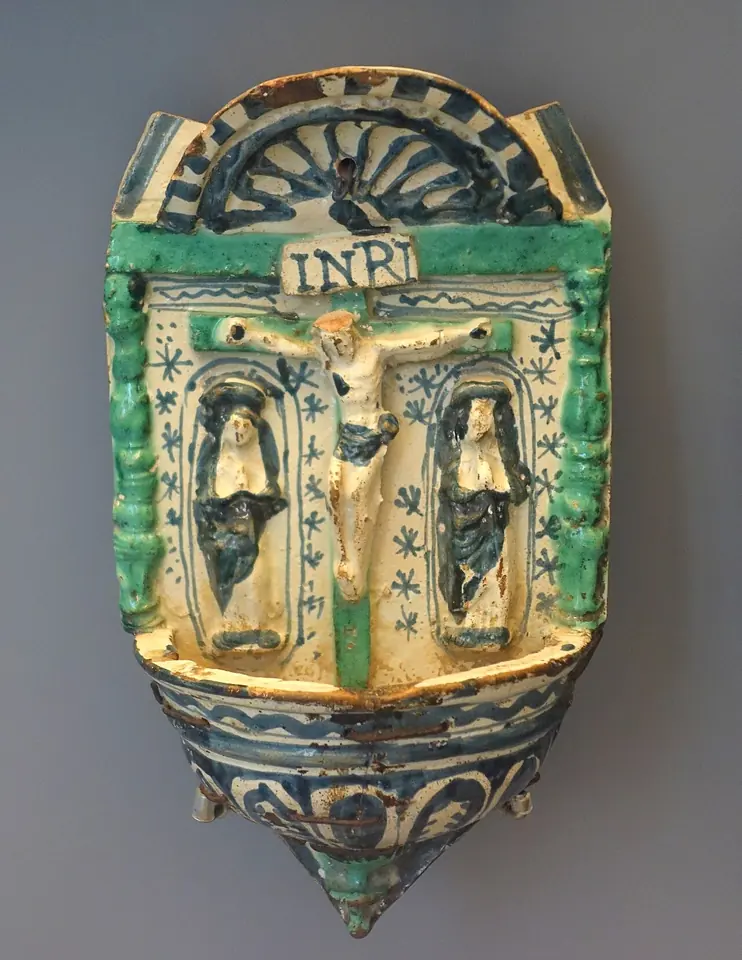 A glazed ceramic wall font in green, blue and yellow, with a small basin below and a relief of Christ on the cross under the letters INRI, flanked by Mary and St. John in arched niches