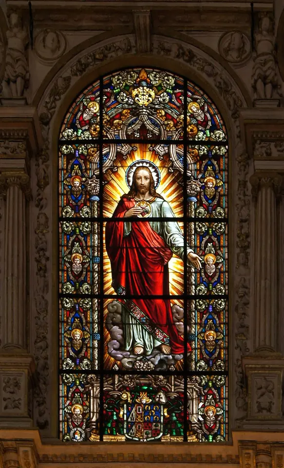 Tall arched stained-glass window of Christ in a red robe and white tunic, his heart shown on his chest in a blaze of golden light, framed by blue and gold ornament