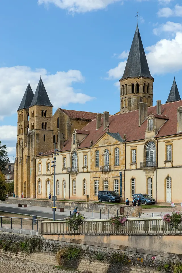 Basílica románica de altas torres cuadradas y, junto a ella, un largo edificio claro, vista desde el otro lado de un río en un día de sol