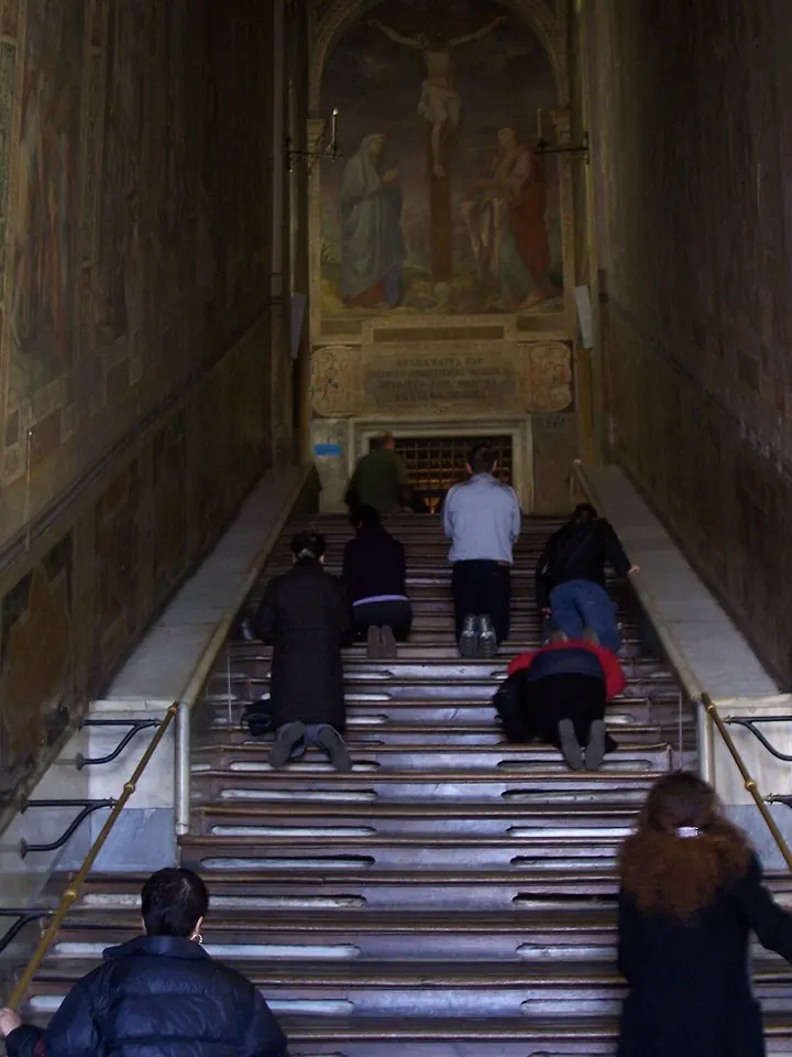 Pilgrims kneeling on a long wood-covered staircase, climbing toward a painting of the crucifixion at the top of a narrow frescoed hall