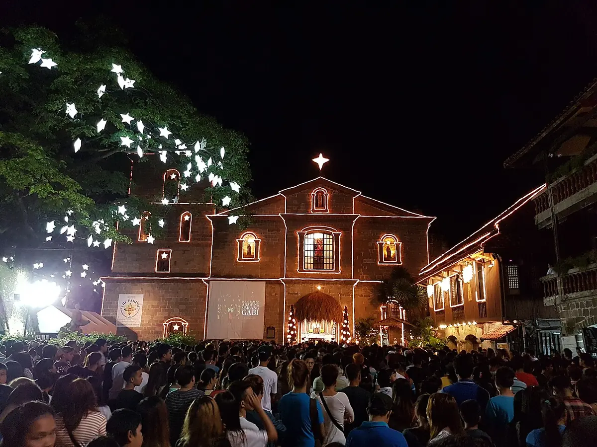 A crowd fills the churchyard of an old brick church at dawn, under trees hung with white star-shaped lights