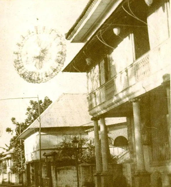 Old sepia photograph of a large round lantern hung high over a street of two-story houses with wooden balconies