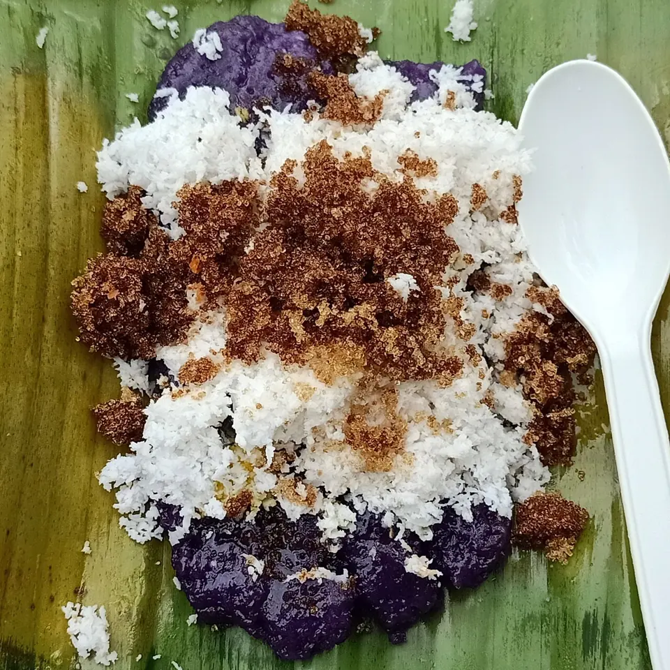 Purple sticky rice cakes on a banana leaf, covered with grated white coconut and brown muscovado sugar, with a plastic spoon