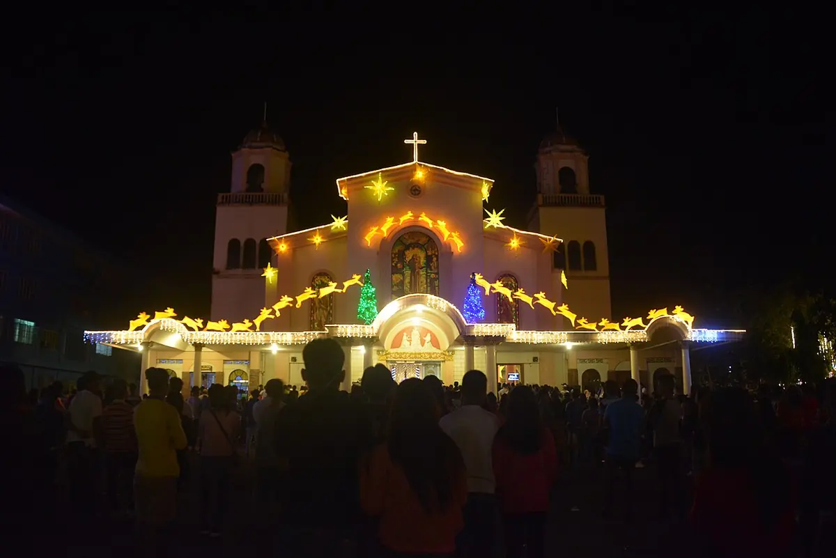 A church facade outlined in Christmas lights at night with a crowd standing in the dark plaza in front of it