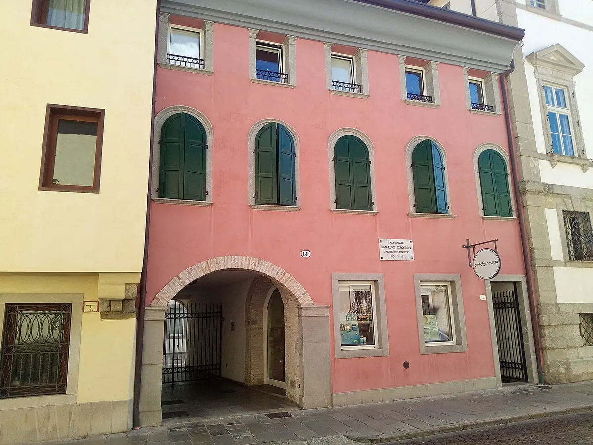 Pink stucco townhouse in Udine with green shutters, an arched carriage entrance and a small memorial plaque beside the door