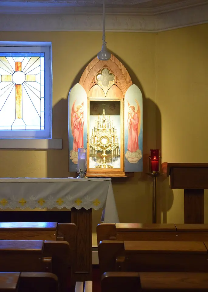 Una pequeña capilla con bancas de madera frente a un altar donde una custodia dorada con el Santísimo Sacramento está en un nicho tallado entre dos ángeles pintados, con una vela roja encendida