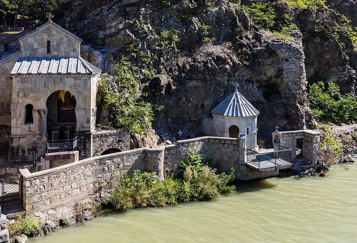 A small stone chapel with a domed kiosk beside it, built against rocky cliffs on the green waters of the Mtkvari River