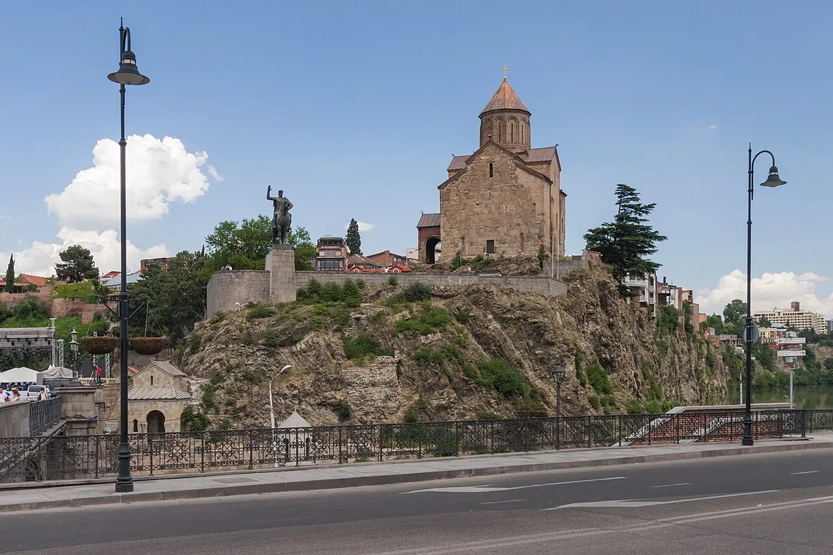 A stone church with a conical dome on a rocky cliff above a road and river in Tbilisi, with an equestrian statue beside it