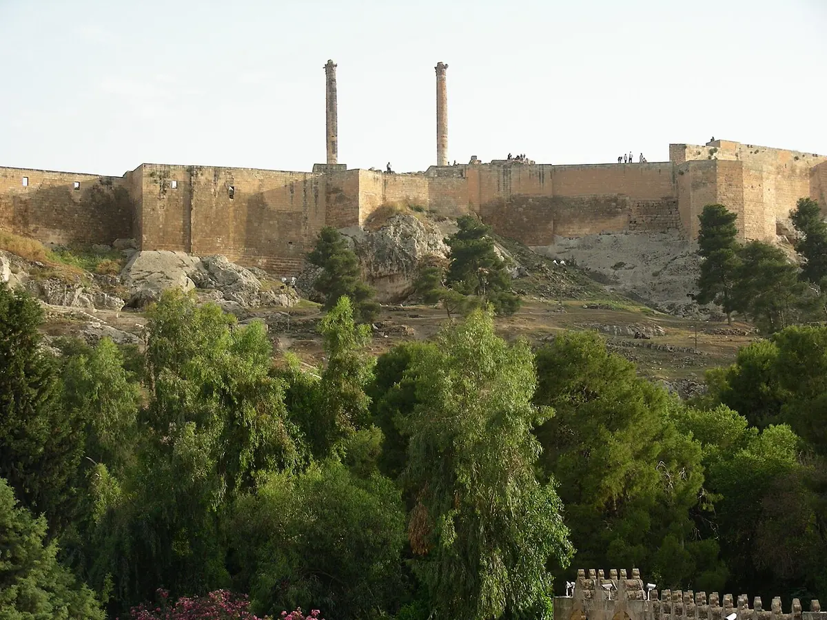 The long stone walls of a hilltop citadel with two tall ancient columns rising above green trees