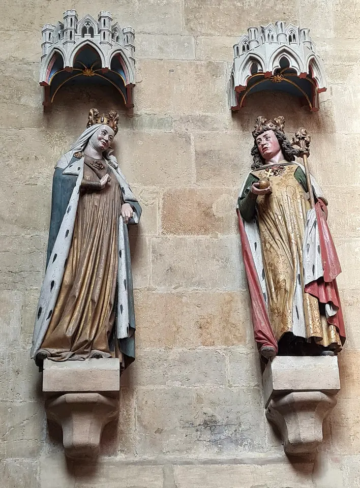 Two painted Gothic stone statues on a cathedral wall, a crowned empress in a white veil and ermine cloak beside a crowned emperor
